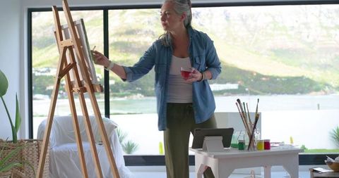 Mature woman painting in bright lakeside home studio with easel, brushes and tablet