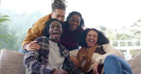 Laughing diverse friends relaxing on sunlit living room sofa, cozy casual gathering