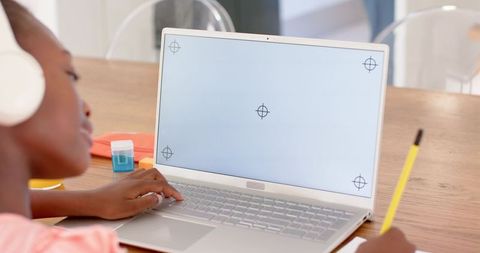 African teen studying at laptop wearing headphones and writing in notebook at home desk