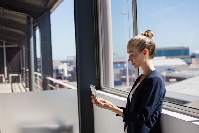 Businesswoman standing by a large window in a modern office, using a digital tablet. She appears focused and professional, dressed in business attire. This image can be used for business, technology, and professional themes, illustrating modern workplace environments, corporate communication, and the use of digital devices in business settings.