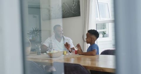 African american father and son painting together at sunlit dining table, creative bonding