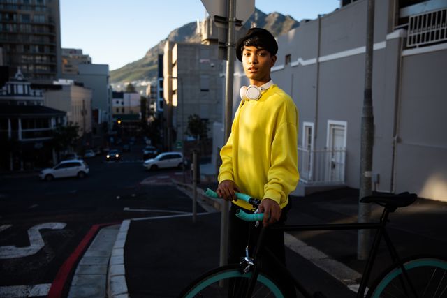 Side view of a fashionable biracial transgender in the street, walking and holding a bike, looking at the camera, with headphones on a neck, waiting to cross a street