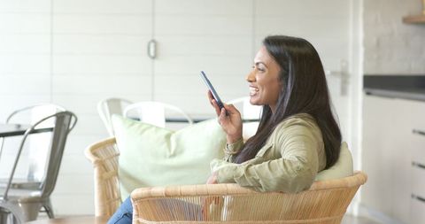 Mid-adult indian woman speaking into smartphone while relaxing in rattan lounge chair