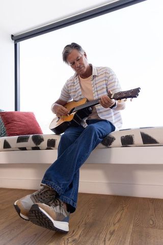 Mature man playing acoustic guitar on sunlit window bench in cozy living room