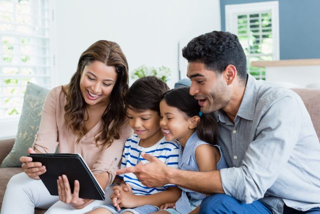 A family of four, consisting of parents and two children, is sitting on a couch in the living room while using a digital tablet. Everyone appears engaged and happy. This image can be used for content related to family bonding, technology use in households, parenting tips, or lifestyle articles.