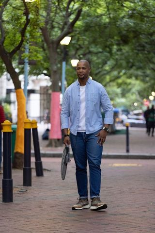 African man walking on tree-lined urban path holding folded umbrella and checking wrist device