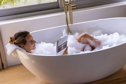Woman relaxing in bubble bath reading book by window in freestanding bathtub