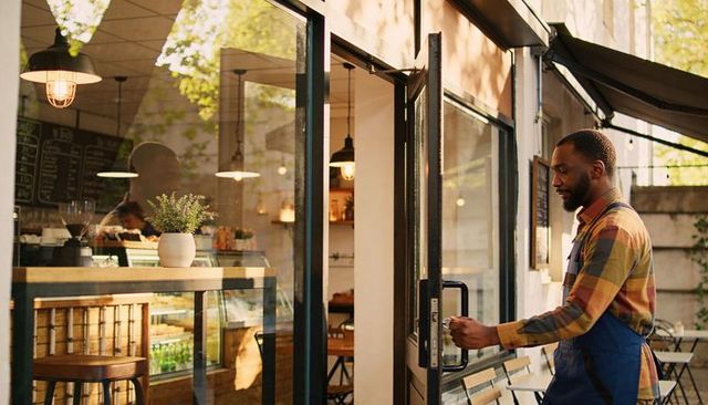 Barista opening glass door at sunny street cafe with pastry counter and outdoor seating