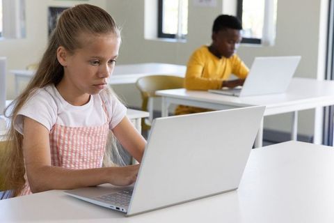Focused girl using laptop in bright classroom with diverse classmates learning on devices