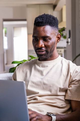 African american man working on laptop with smartwatch in bright modern living room