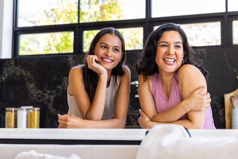 Smiling diverse women laughing and leaning on sunlit modern kitchen counter, home lifestyle