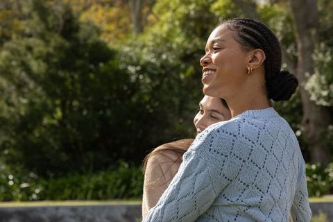Sunlit diverse friends embracing and smiling in garden wearing blue sweater and gold hoops