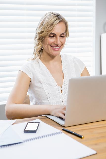 Woman sitting at a wooden desk, using a laptop and smiling. Ideal for depicting remote work, home office setups, freelance lifestyle, and business productivity. Suitable for articles, blogs, and advertisements about remote working, home offices, and women in business.