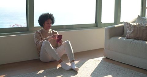 Man relaxing by ocean window using smartphone on sunlit hardwood floor, minimal lifestyle