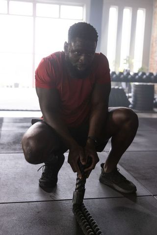 African american man squatting and gripping battle rope in intense gym strength training