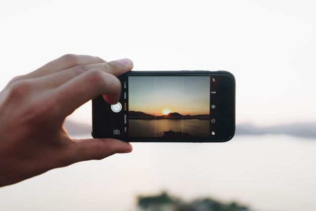 A hand capturing the scenic seaside sunset with a smartphone. This photo can be ideal for illustrating modern travel experiences, leisure activities, or the use of technology in capturing nature's beauty. Perfect for blogs, travel agencies and electronic gadget promotions.
