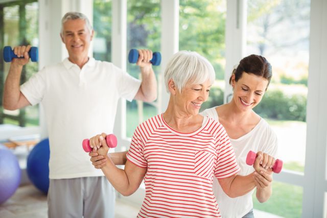 Female trainer assisting senior couple in performing exercise using dumbbells at home. Bright indoor environment with large windows. Ideal for fitness and wellness articles, senior health promotions, and personal training services targeting elderly clients.