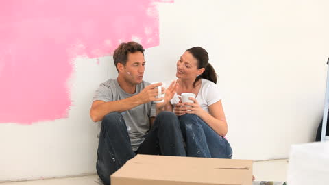 Couple taking a moment to relax and enjoy coffee while painting and renovating their home. This video is ideal for depicting teamwork, bonding during a DIY home project, or advertising lifestyle products focusing on home improvement activities.