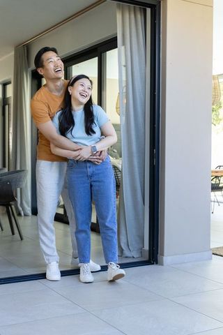 Asian couple hugging and laughing in modern home by sliding glass door, smartwatch, romance