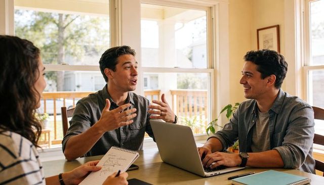Man leading informal home meeting gesturing at sunlit dining table with laptop