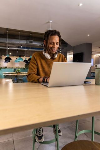 African American male working on laptop in modern cafe wearing headphones and smiling