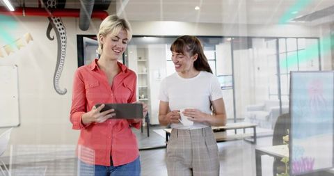 Two female colleagues collaborating with tablet and coffee in modern glass office space