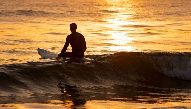 Surfer silhouette sitting on surfboard at golden sunset ocean reflection
