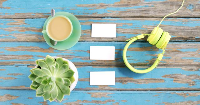 A cup of coffee, headphones, and a succulent plant are arranged on a rustic blue wooden table, with blank business cards providing copy space. It suggests a creative or casual business setting, ideal for brainstorming or relaxation.