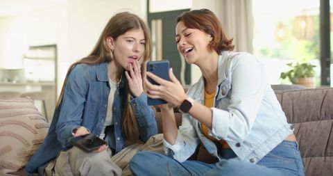 Diverse women laughing while sharing smartphone video on sofa in cozy sunlit living room