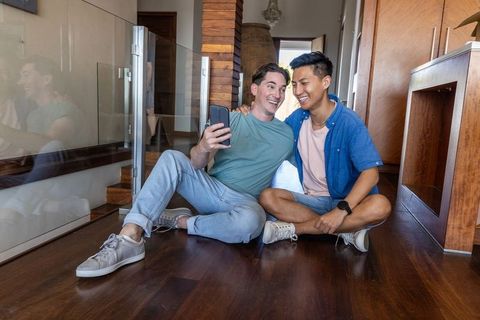 Two men sitting on hardwood floor taking selfie by glass railing in modern home