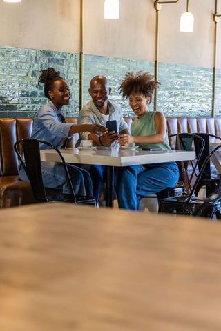 African american friends laughing and sharing smartphone while sitting in cafe booth