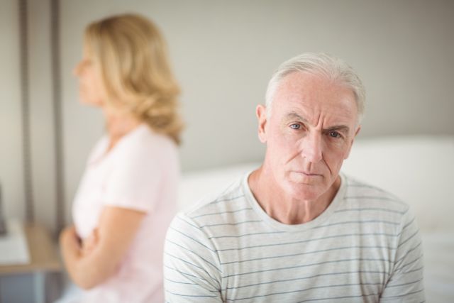Senior man sitting on bed looking worried while woman sits in background. Useful for topics on aging, relationships, emotional health, and senior lifestyle. Ideal for articles, blogs, and advertisements focusing on elderly care, mental health, and family dynamics.