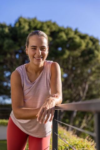 Sunlit fitness portrait of smiling woman leaning on balcony railing in pink activewear