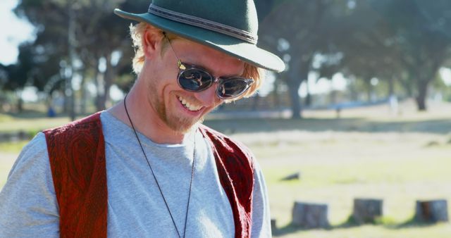 Young man enjoying a sunny day outdoors, wearing a hat and sunglasses. Ideal for content related to relaxation, happiness, leisure activities, fashion, and outdoor lifestyle. Perfect for advertising, blogs, and social media posts focused on youthful energy and carefree living.