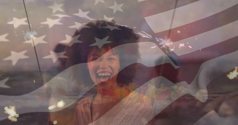 Laughing woman holding sparkler on beach at dusk with american flag overlay, patriotic