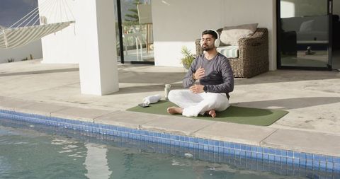 Indian man meditating by pool on yoga mat with headphones sunlit outdoor mindfulness
