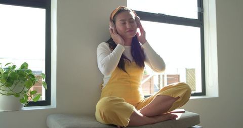 Mid-adult asian woman relaxing and listening to music on headphones in sunlit window seat