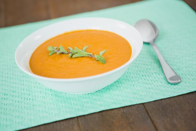 Bowl of creamy pumpkin soup garnished with fresh herbs, placed on a green tablecloth with a spoon beside it. Ideal for use in food blogs, recipe websites, healthy eating promotions, and autumn-themed content.