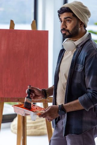 Young artist standing painting red canvas at easel wearing beanie and headphones