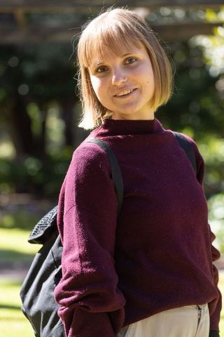 Young woman standing, smiling in sunlit garden wearing burgundy sweater and black backpack