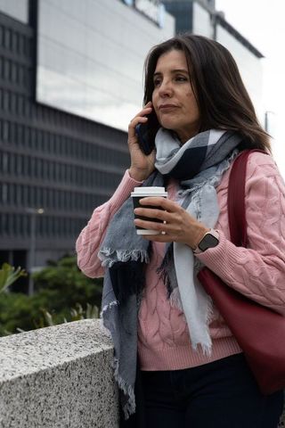 Middle-aged woman talking on phone holding coffee wearing scarf and smartwatch in city commute