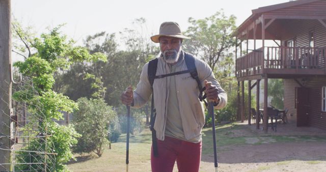 A senior man trekking near a wooden cabin in a rural setting, using walking poles. The man is dressed in outdoor gear and wearing a hat, highlighting his active and adventurous lifestyle. This image can be used for promoting healthy living, outdoor activities, retirement lifestyles, and travel options for seniors.