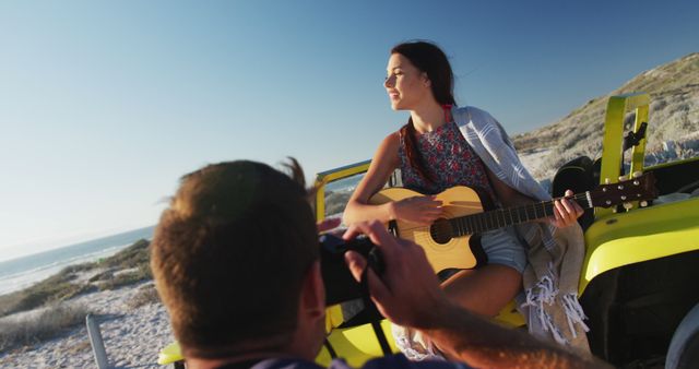 Caucasian man taking pictures of woman sitting in beach buggy by the sea playing guitar. beach stop off on summer holiday road trip.