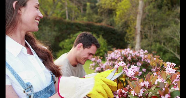 A couple is happily engaged in gardening activities, trimming flowers in a lush green backyard. Both are wearing protective gloves while focusing on their tasks, displaying teamwork and a love for nature. Perfect visuals for themes related to gardening, home improvement, outdoor activities, and bonding time.