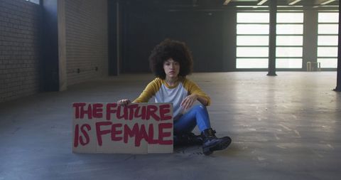 This portrays a strong and confident woman sitting in a parking garage, holding a placard advocating for gender equality and female empowerment. Ideal for use in campaigns related to women's rights, feminism, activism, diversity representation, or discussions on gender expression and identity.