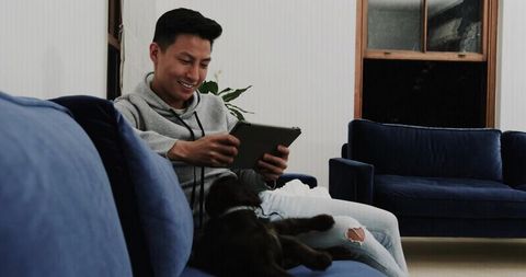 Asian man relaxing on navy velvet sofa using tablet with dog, modern cozy living room