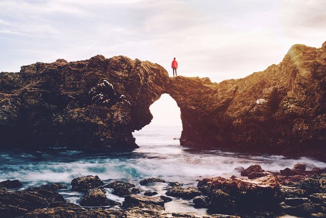 Person standing on a cliff with a natural arch overlooking the ocean at sunset. Waves crash against the rocky shoreline as the sunlight creates a warm ambiance. Ideal for use in travel advertisements, adventure magazines, or inspirational posters focusing on nature and solitude.