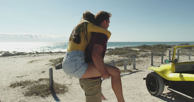 An affectionate couple is spending time on a sunny beach. The woman, wearing a yellow shirt and denim shorts, is getting a piggyback ride from the man. The scene includes the vast ocean, sandy shore, and a yellow dune buggy, suggesting a day filled with fun and adventure. Ideal for romantic getaway promotions, travel and tourism advertising, lifestyle blogs, and vacation planning resources.