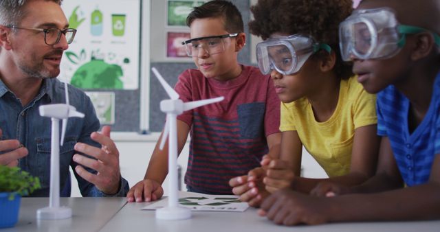 Teacher is explaining the mechanics of wind turbine models to a group of diverse students. The students are wearing safety goggles and attentively listening, indicating engagement in a science or renewable energy project. This image is suitable for use in educational materials, websites promoting STEM education, renewable energy initiatives, or classroom activities.