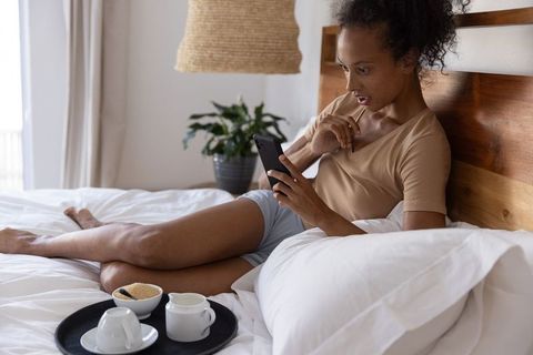 Surprised african american woman reclining on bed holding smartphone for morning relaxation